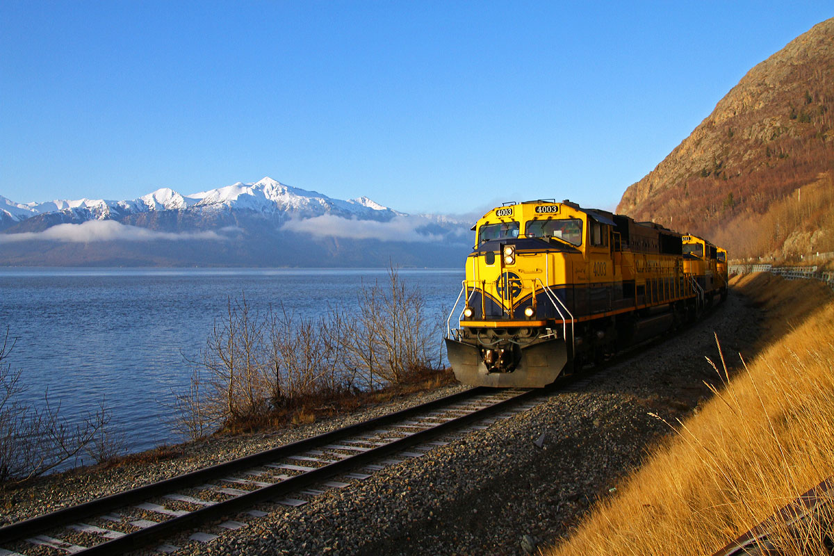 An Alaska RR empty lumber trains rolls along Turnagain Arm, in the background are the Kenai Mountains.