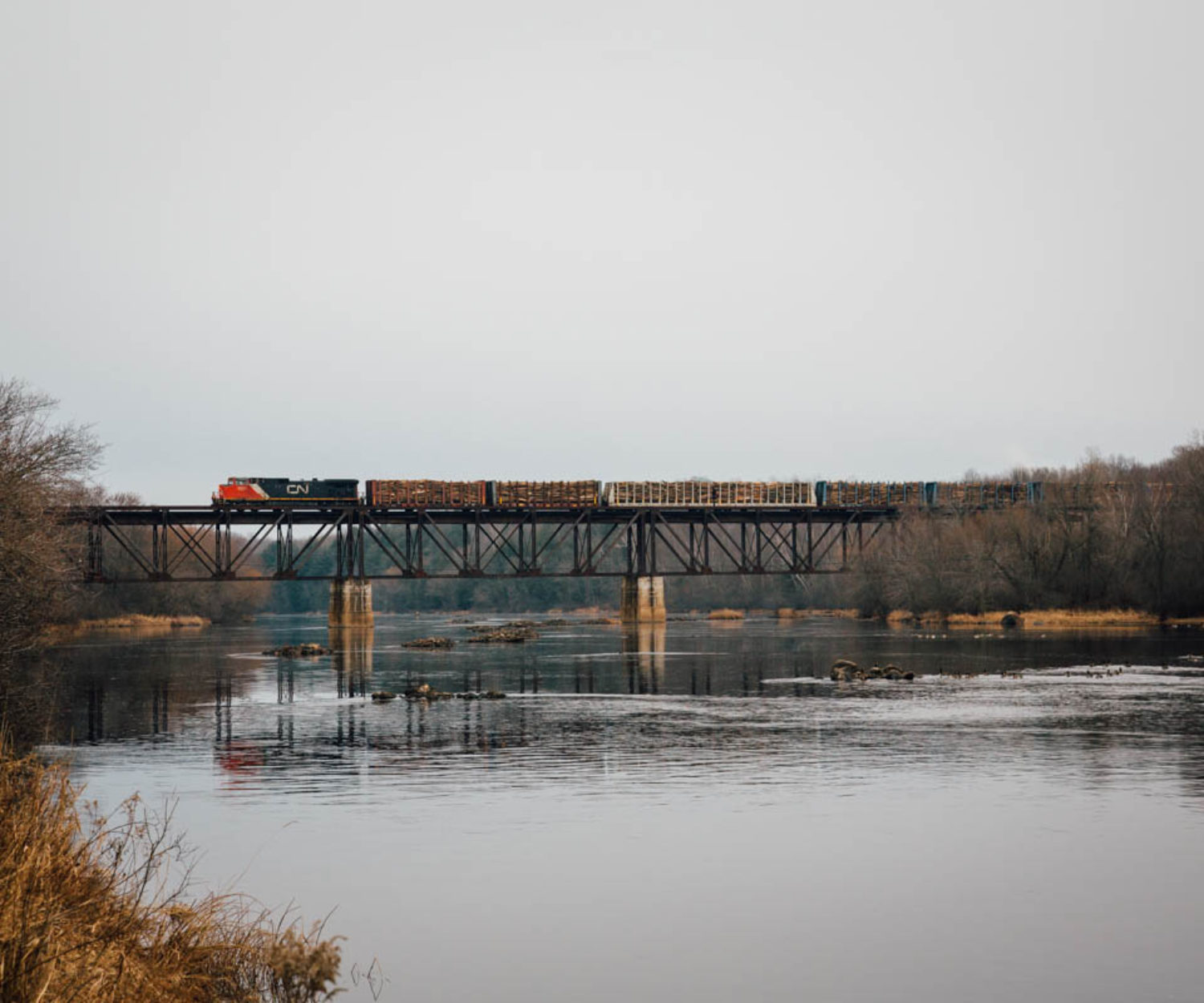 A lone Canadian National locomotive pushes a large cut of bulkhead flat cars loaded with logs over the Flambeau River. They will cut off the three rear cars at Besse Lumber Company just south of the river before heading back north on the Superior Sub. 