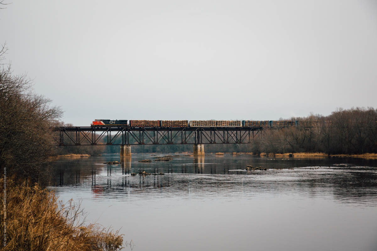 A lone Canadian National locomotive pushes a large cut of bulkhead flat cars loaded with logs over the Flambeau River. They will cut off the three rear cars at Besse Lumber Company just south of the river before heading back north on the Superior Sub. 