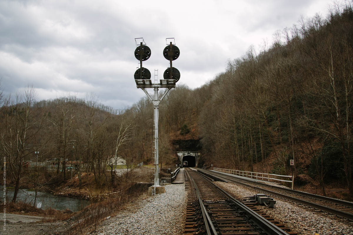 The Norfolk and Western Railway's coal hauling right of way once followed the course of the Tug River between Welch and Davy. In 1903, the railroad started a major rebuilding project to straighten out portions of the Pocahontas Division and prepare for future double tracking. Just a year later, two tunnels were constructed roughly a mile norwest of Welch, known as Hemphill. Hemphill originated as a name of a siding, likely named after Secretary of the N&W, A.J. Hemphill in 1894, which eventually evolved into a community. The two tunnels named Hemphill Tunnel No. 1 and No. 2 respectively were cut through the Appalachian Plateaus Physiographic Province, a region characterized by deeply incised plateaus underlain by flat-lying sedimentary rock. The railroad grade was raised during this realignment to avoid the common flooding of the Tug Fork which it crosses three times in this short stretch. The realignment shaved off 2.4 miles from the previous wandering Tug Fork route. Double tracking was completed here in 1909. The N&W CPL Signal can be seen by trains exiting the west portal of tunnel no. 2 that guards Farm, a well known railroad location where a large coaling tower still stands. Today, helpers wait assignments to shove heavily loaded coal trains east up the steep steady grade.